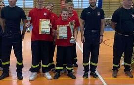 Zdjęcia przedstawiają uczestnik&oacute;w w strojach sportowych i strażackich podczas rywalizacji na hali sportowej, m.in. dynamiczne konkurencje z wężami strażackimi, uroczystą zbi&oacute;rkę wszystkich drużyn, a także momenty wręczania puchar&oacute;w, dyplom&oacute;w oraz nagr&oacute;d indywidualnych przez zaproszonych gości i przedstawicieli straży pożarnej.