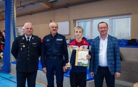 Zdjęcia przedstawiają uczestnik&oacute;w w strojach sportowych i strażackich podczas rywalizacji na hali sportowej, m.in. dynamiczne konkurencje z wężami strażackimi, uroczystą zbi&oacute;rkę wszystkich drużyn, a także momenty wręczania puchar&oacute;w, dyplom&oacute;w oraz nagr&oacute;d indywidualnych przez zaproszonych gości i przedstawicieli straży pożarnej.