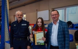 Zdjęcia przedstawiają uczestnik&oacute;w w strojach sportowych i strażackich podczas rywalizacji na hali sportowej, m.in. dynamiczne konkurencje z wężami strażackimi, uroczystą zbi&oacute;rkę wszystkich drużyn, a także momenty wręczania puchar&oacute;w, dyplom&oacute;w oraz nagr&oacute;d indywidualnych przez zaproszonych gości i przedstawicieli straży pożarnej.