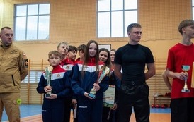 Zdjęcia przedstawiają uczestnik&oacute;w w strojach sportowych i strażackich podczas rywalizacji na hali sportowej, m.in. dynamiczne konkurencje z wężami strażackimi, uroczystą zbi&oacute;rkę wszystkich drużyn, a także momenty wręczania puchar&oacute;w, dyplom&oacute;w oraz nagr&oacute;d indywidualnych przez zaproszonych gości i przedstawicieli straży pożarnej.