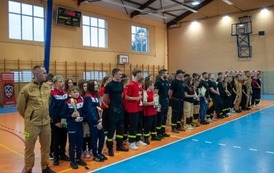 Zdjęcia przedstawiają uczestnik&oacute;w w strojach sportowych i strażackich podczas rywalizacji na hali sportowej, m.in. dynamiczne konkurencje z wężami strażackimi, uroczystą zbi&oacute;rkę wszystkich drużyn, a także momenty wręczania puchar&oacute;w, dyplom&oacute;w oraz nagr&oacute;d indywidualnych przez zaproszonych gości i przedstawicieli straży pożarnej.