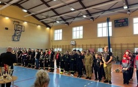 Zdjęcia przedstawiają uczestnik&oacute;w w strojach sportowych i strażackich podczas rywalizacji na hali sportowej, m.in. dynamiczne konkurencje z wężami strażackimi, uroczystą zbi&oacute;rkę wszystkich drużyn, a także momenty wręczania puchar&oacute;w, dyplom&oacute;w oraz nagr&oacute;d indywidualnych przez zaproszonych gości i przedstawicieli straży pożarnej.