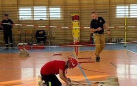 Zdjęcia przedstawiają uczestnik&oacute;w w strojach sportowych i strażackich podczas rywalizacji na hali sportowej, m.in. dynamiczne konkurencje z wężami strażackimi, uroczystą zbi&oacute;rkę wszystkich drużyn, a także momenty wręczania puchar&oacute;w, dyplom&oacute;w oraz nagr&oacute;d indywidualnych przez zaproszonych gości i przedstawicieli straży pożarnej.