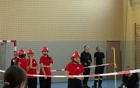 Zdjęcia przedstawiają uczestnik&oacute;w w strojach sportowych i strażackich podczas rywalizacji na hali sportowej, m.in. dynamiczne konkurencje z wężami strażackimi, uroczystą zbi&oacute;rkę wszystkich drużyn, a także momenty wręczania puchar&oacute;w, dyplom&oacute;w oraz nagr&oacute;d indywidualnych przez zaproszonych gości i przedstawicieli straży pożarnej.