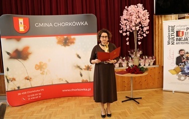 Kobieta w ciemnobrązowej sukience i okularach stoi przy mikrofonie, trzymając otwartą czerwoną teczkę i przemawiając do zgromadzonych. W tle znajduje się ciemnoczerwona kurtyna, dekoracyjne drzewko z r&oacute;żowymi kwiatami, st&oacute;ł z bukietem czerwonych r&oacute;ż oraz ścianki reklamowe z napisem &bdquo;Gmina Chork&oacute;wka&rdquo;.