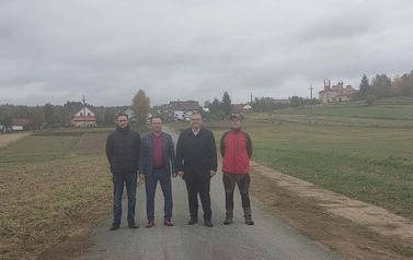 Czterech mężczyzn stoi w rzędzie na utwardzonej drodze gruntowej, patrząc w stronę aparatu. Są ubrani w odzież wierzchnią: od lewej czarna kurtka puchowa, bordowy sweter pod marynarką, czarny płaszcz, czerwono-czarna kurtka sportowa. Tło jest identyczne jak na poprzednim zdjęciu &ndash; wiejska droga prowadząca do oddalonej zabudowy z kościołem na wzg&oacute;rzu, otoczona polami. Niebo jest całkowicie zachmurzone.