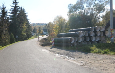 Prace budowlane przy drodze powiatowej polegające na budowie nowego chodnika oraz profilowaniu i umacnianiu row&oacute;w odwadniających. Na zdjęciach widoczne są odcinki drogi z ustawionymi biało-czerwonymi pachołkami ostrzegawczymi, wykopana i wyr&oacute;wnana ziemia pod krawężniki i podbudowę, a także fragmenty row&oacute;w wyłożone ażurowymi płytami betonowymi. W tle widoczne są domy jednorodzinne, zielone oraz jesienne drzewa i znaki drogowe.