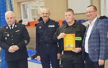 Zdjęcia przedstawiają uczestnik&oacute;w w strojach sportowych i strażackich podczas rywalizacji na hali sportowej, m.in. dynamiczne konkurencje z wężami strażackimi, uroczystą zbi&oacute;rkę wszystkich drużyn, a także momenty wręczania puchar&oacute;w, dyplom&oacute;w oraz nagr&oacute;d indywidualnych przez zaproszonych gości i przedstawicieli straży pożarnej.