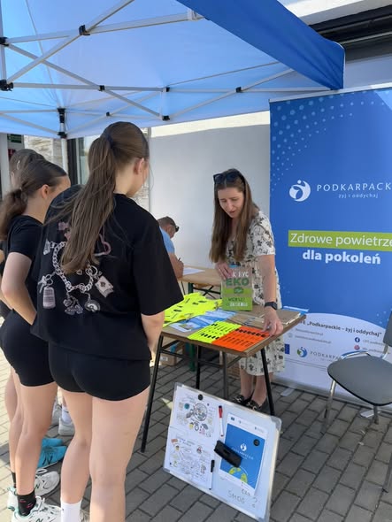 Kobieta stojąca przy stanowisku edukacyjnym rozmawia z&nbsp;grupą nastolatek stojących tyłem do obiektywu. Kobieta trzyma w&nbsp;dłoni zieloną książkę lub&nbsp;ulotkę z&nbsp;napisem &bdquo;Jak być eko&rdquo;. Nad stoiskiem rozłożony jest biało-niebieski namiot, a&nbsp;obok stoi niebieski roll-up z&nbsp;hasłem &bdquo;Zdrowe powietrze dla pokoleń&rdquo;. Na stole promocyjnym leżą odblaski i&nbsp;ulotki, a&nbsp;na ziemi przed nim widoczna jest tablica edukacyjna dotycząca ekologii i&nbsp;smogu.