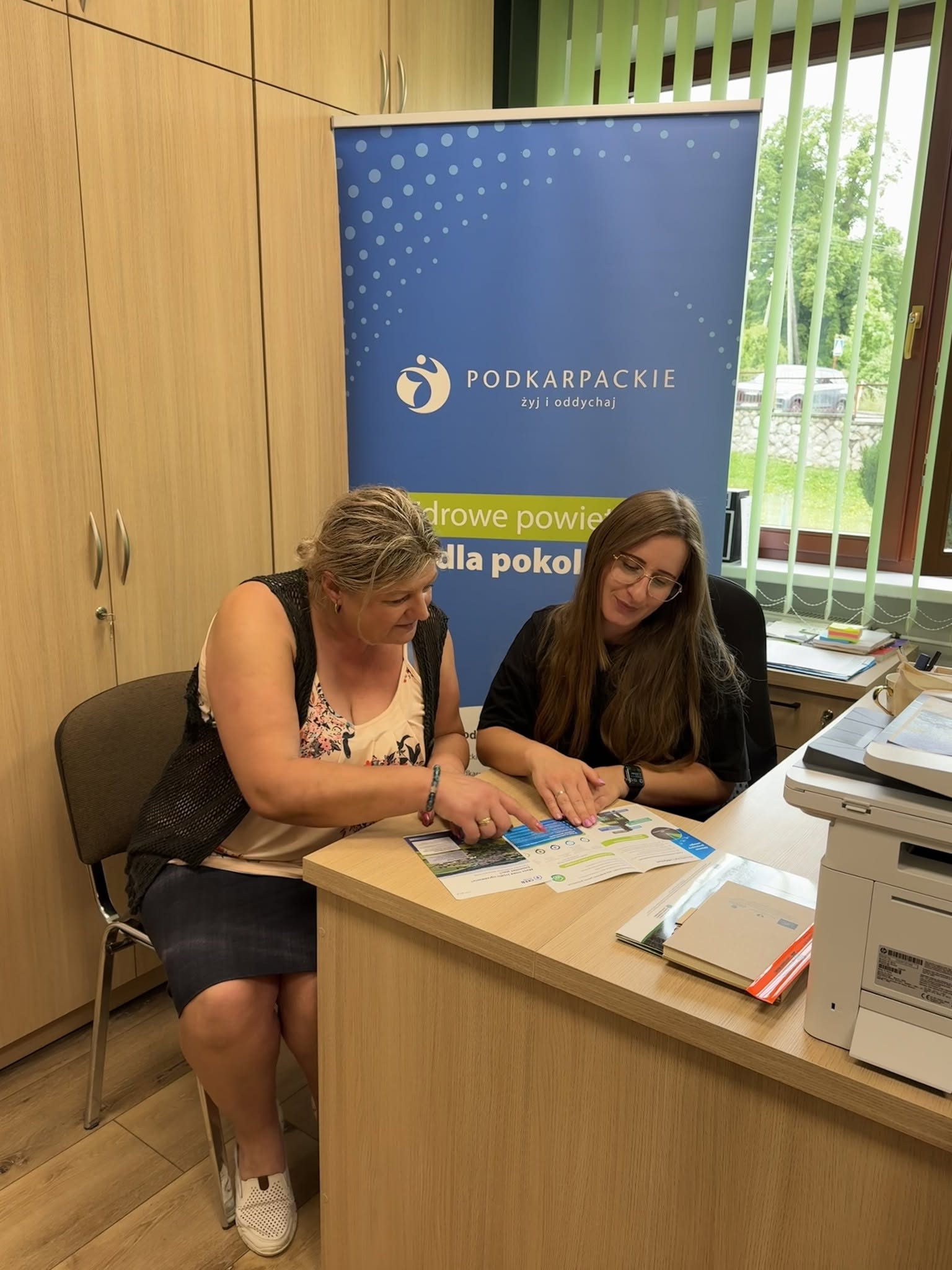 Dwie kobiety siedzą za biurkiem i&nbsp;przeglądają ulotki. 