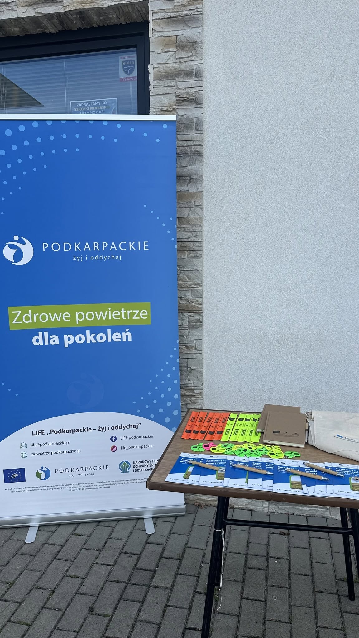 Galeria 3 zdjęć przedstawiająca plenerowe stoisko informacyjno-promocyjne projektu LIFE &bdquo;Podkarpackie &ndash; żyj i&nbsp;oddychaj&rdquo;. Na tle niebieskiego banneru z&nbsp;hasłem &bdquo;Zdrowe powietrze dla pokoleń&rdquo; stoi stolik z&nbsp;materiałami edukacyjnymi i&nbsp;gadżetami: ulotkami o&nbsp;dotacjach na&nbsp;termomodernizację, ekologicznymi długopisami, notesami, odblaskami i&nbsp;materiałowymi torbami. Na jednym ze zdjęć młoda dziewczynka ogląda gadżety przy stoliku w&nbsp;asyście dorosłej kobiety.