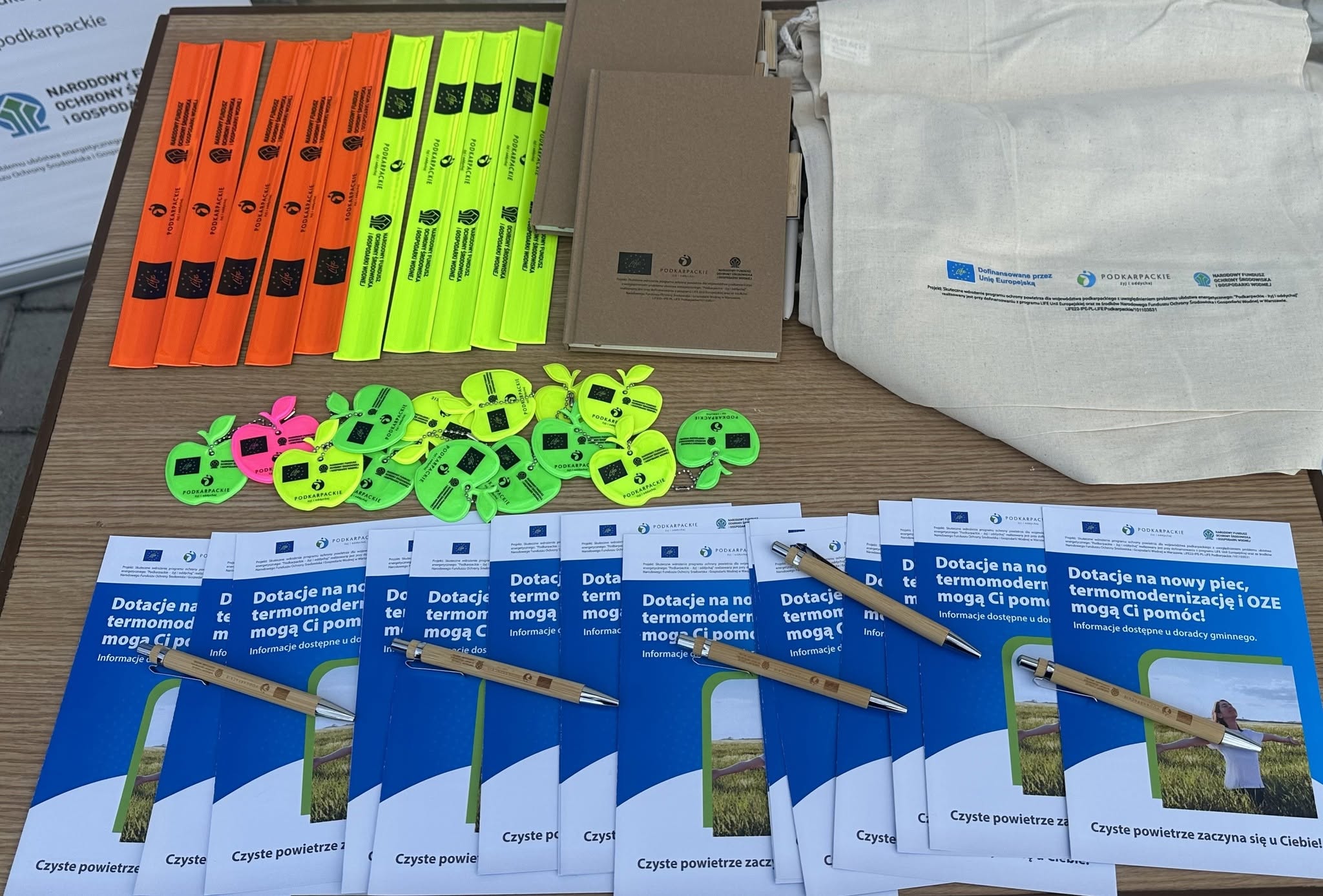 Galeria 3 zdjęć przedstawiająca plenerowe stoisko informacyjno-promocyjne projektu LIFE &bdquo;Podkarpackie &ndash; żyj i&nbsp;oddychaj&rdquo;. Na tle niebieskiego banneru z&nbsp;hasłem &bdquo;Zdrowe powietrze dla pokoleń&rdquo; stoi stolik z&nbsp;materiałami edukacyjnymi i&nbsp;gadżetami: ulotkami o&nbsp;dotacjach na&nbsp;termomodernizację, ekologicznymi długopisami, notesami, odblaskami i&nbsp;materiałowymi torbami. Na jednym ze zdjęć młoda dziewczynka ogląda gadżety przy stoliku w&nbsp;asyście dorosłej kobiety.