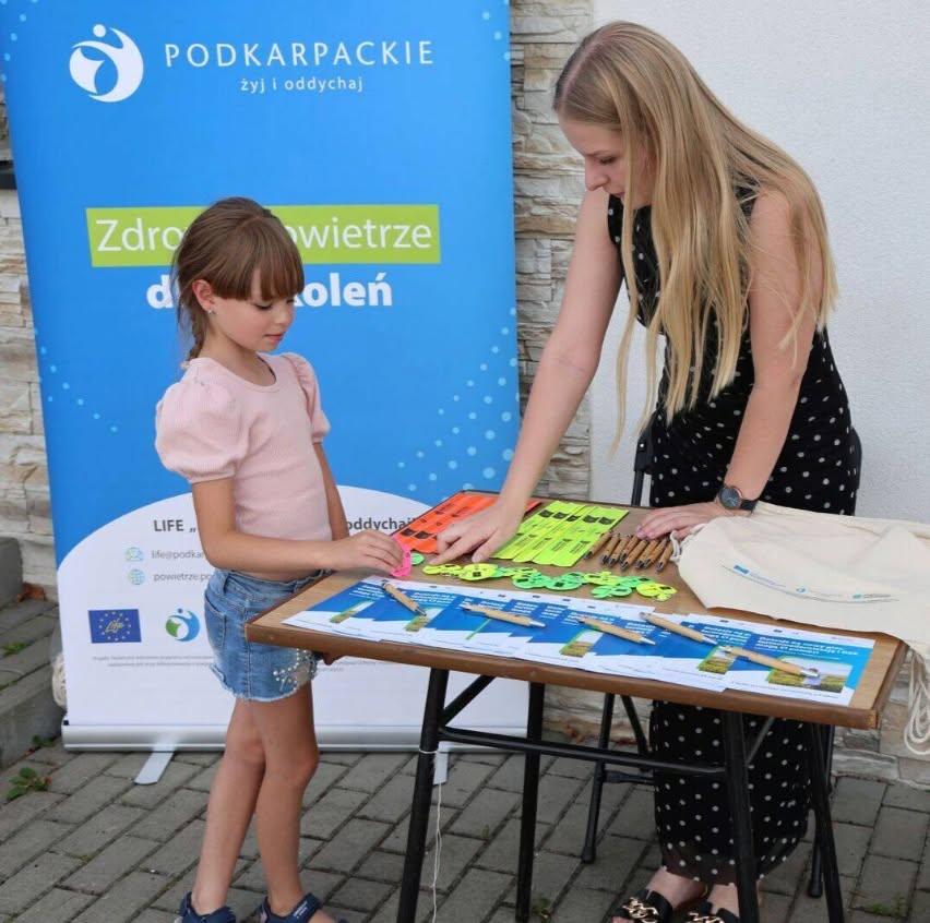 Galeria 3 zdjęć przedstawiająca plenerowe stoisko informacyjno-promocyjne projektu LIFE &bdquo;Podkarpackie &ndash; żyj i&nbsp;oddychaj&rdquo;. Na tle niebieskiego banneru z&nbsp;hasłem &bdquo;Zdrowe powietrze dla pokoleń&rdquo; stoi stolik z&nbsp;materiałami edukacyjnymi i&nbsp;gadżetami: ulotkami o&nbsp;dotacjach na&nbsp;termomodernizację, ekologicznymi długopisami, notesami, odblaskami i&nbsp;materiałowymi torbami. Na jednym ze zdjęć młoda dziewczynka ogląda gadżety przy stoliku w&nbsp;asyście dorosłej kobiety.