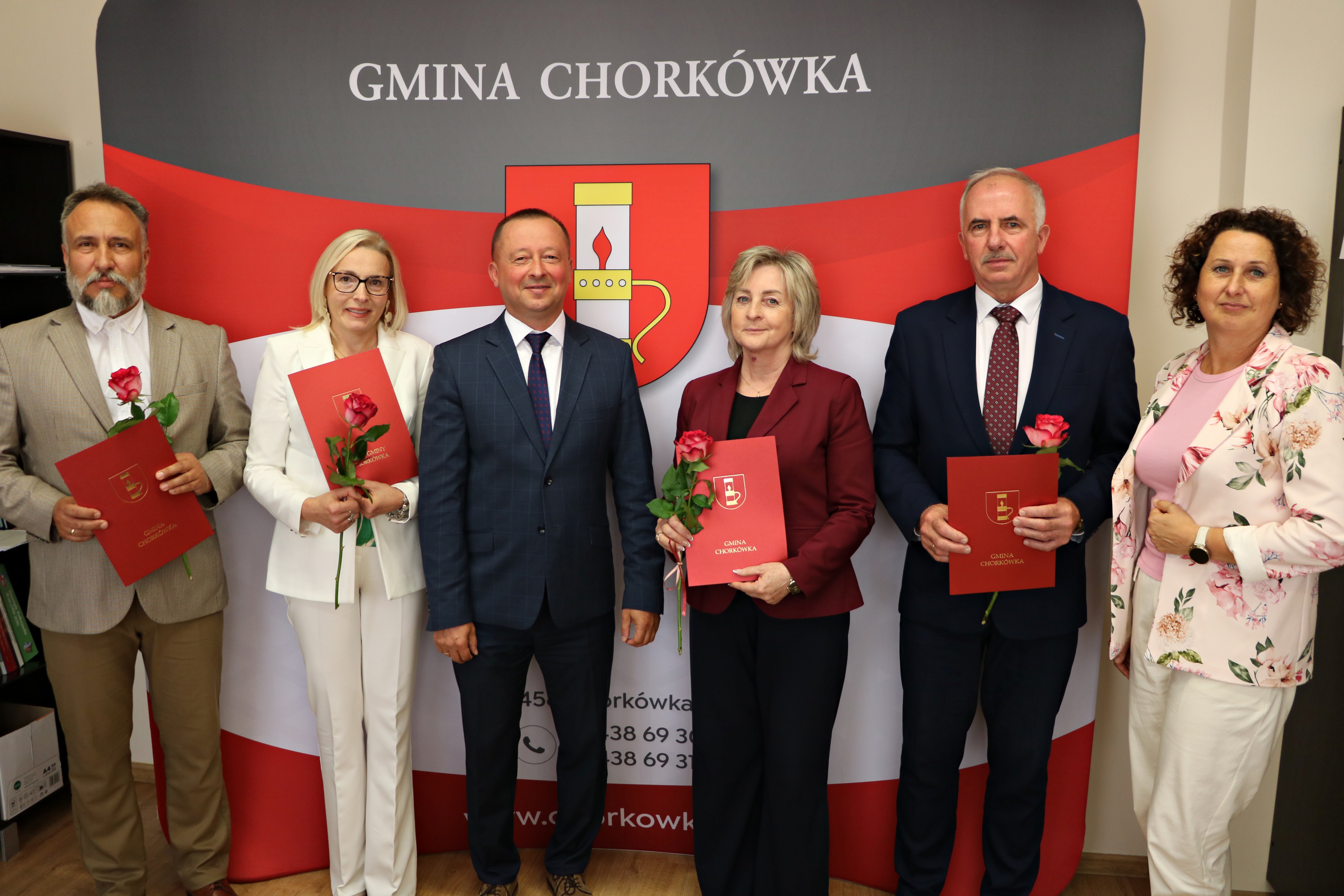 Fotografia grupowa sześciu os&oacute;b, stojących przed tą samą biało-czerwoną ścianką reklamową Gminy Chork&oacute;wka, co na&nbsp;zdjęciu image_0.png. Skład grupy jest identyczny jak na&nbsp;pierwszym zdjęciu (W&oacute;jt w&nbsp;centrum, czworo dyrektor&oacute;w z&nbsp;r&oacute;żami i&nbsp;teczkami), jednak&nbsp;skrajnie po prawej stronie dołączyła sz&oacute;sta osoba: kobieta o&nbsp;kręconych, ciemnych włosach, ubrana w&nbsp;białą marynarkę w&nbsp;kwiatowe wzory. Wszyscy pozują przodem do aparatu w&nbsp;oficjalnych, uśmiechniętych pozach.
