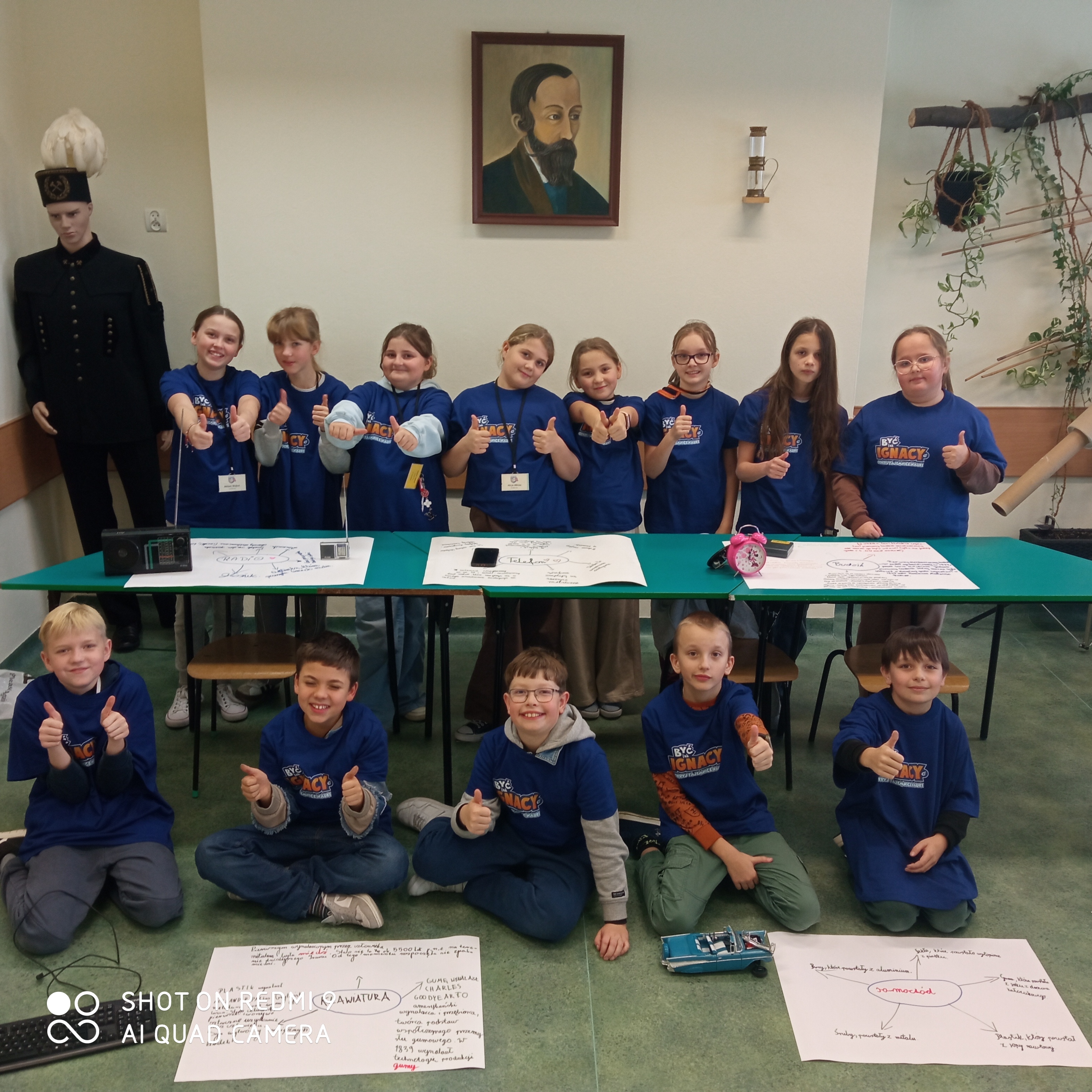 Na zdjęciu znajduje się grupa trzynastu uśmiechniętych dzieci w&nbsp;wieku szkolnym, pozujących w&nbsp;sali edukacyjnej. Dzieci mają na&nbsp;sobie pasujące ciemnoniebieskie koszulki z&nbsp;pomarańczowo-białym logo &bdquo;Być jak Ignacy&rdquo;. Większość z&nbsp;nich pokazuje gest kciuka w&nbsp;g&oacute;rę.  Układ grupy:  W g&oacute;rnym rzędzie: Osiem dziewczynek stoi za długim, zielonym stołem.  W dolnym rzędzie: Pięciu chłopc&oacute;w siedzi na&nbsp;podłodze przed stołem.  Otoczenie i&nbsp;rekwizyty:  Projekty edukacyjne: Na stole i&nbsp;na podłodze rozłożone są duże białe arkusze papieru z&nbsp;ręcznie narysowanymi mapami myśli i&nbsp;rysunkami (m.in. radia, telefonu, klawiatury i&nbsp;samochodu).  Przedmioty na&nbsp;stole: Widoczne są dwa czarne radia tranzystorowe z&nbsp;antenami oraz&nbsp;r&oacute;żowy budzik. Na podłodze przed jednym z&nbsp;chłopc&oacute;w stoi model niebieskiego samochodu.  Tło: Na jasnej ścianie w&nbsp;centralnym punkcie wisi portret mężczyzny z&nbsp;brodą (prawdopodobnie Ignacego Łukasiewicza). Po lewej stronie stoi manekin w&nbsp;czarnym mundurze g&oacute;rniczym z&nbsp;charakterystyczną czapką z&nbsp;białym pi&oacute;ropuszem. Po prawej stronie widać fragmenty dekoracji z&nbsp;roślinami i&nbsp;konstrukcjami z&nbsp;patyk&oacute;w.