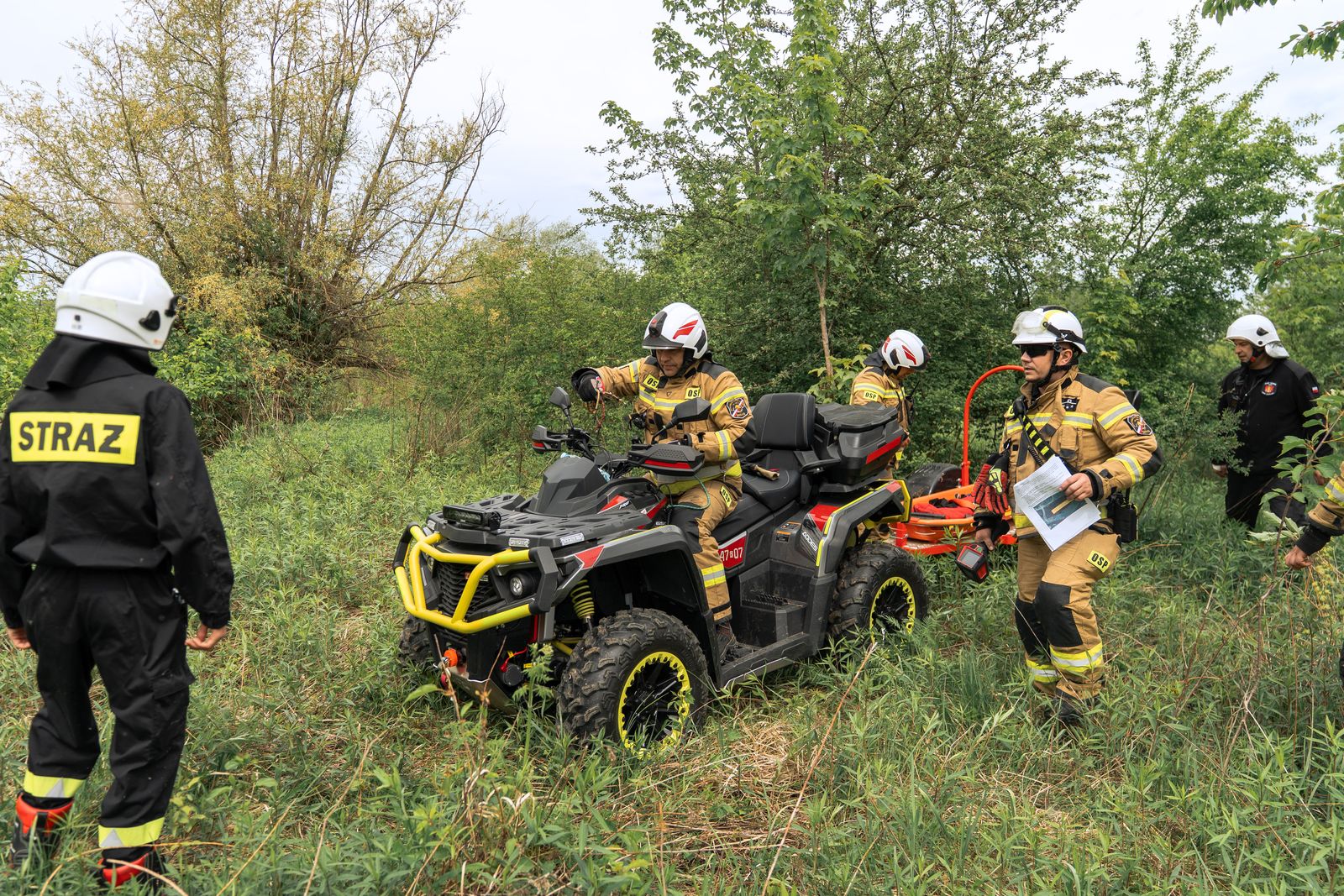 Strażacy w&nbsp;pełnym ekwipunku ochronnym i&nbsp;kaskach przeprowadzają akcję lub&nbsp;ćwiczenia w&nbsp;gęstych zaroślach z&nbsp;użyciem czarnego pojazdu typu quad (ATV).