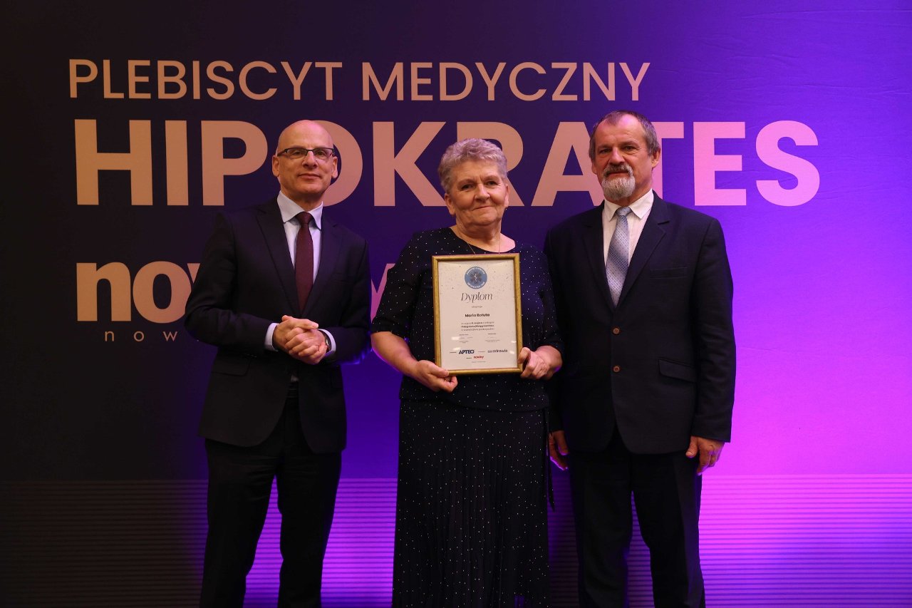 Trzy elegancko ubrane osoby stoją na&nbsp;scenie podczas uroczystej gali medycznej. Kobieta pośrodku trzyma teczkę z&nbsp;dyplomem, a&nbsp;po jej bokach stoją dwaj mężczyźni w&nbsp;garniturach. W tle widoczny jest baner z&nbsp;napisem &bdquo;Plebiscyt Medyczny HIPOKRATES&rdquo; oraz&nbsp;logo &bdquo;nowiny&rdquo;. Na pierwszym planie fotograf robi zdjęcie laureatom.