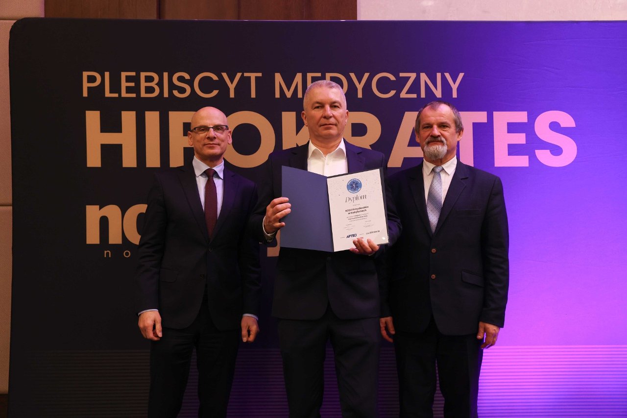 Trzech mężczyzn w&nbsp;formalnych garniturach pozuje do zdjęcia na&nbsp;tle banera &bdquo;Plebiscyt Medyczny HIPOKRATES&rdquo;. Mężczyzna stojący pośrodku trzyma otwarty dyplom skierowany w&nbsp;stronę obiektywu. Całość oświetlona jest eleganckim, scenicznych światłem w&nbsp;odcieniach fioletu i&nbsp;granatu.