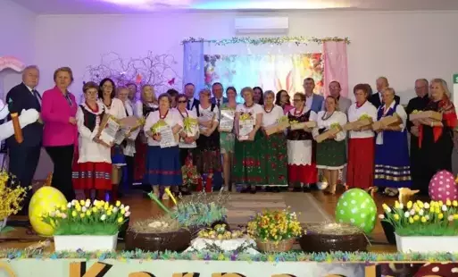 Na zdjęciu znajduje się duża grupa osób stojących na scenie, prawdopodobnie uczestników wydarzenia kulturalnego lub konkursu. W tle sceny widoczna jest dekoracja nawiązująca do świąt wielkanocnych – kolorowe kwiaty, gałązki, pisanki, ozdobny zajączek i ba