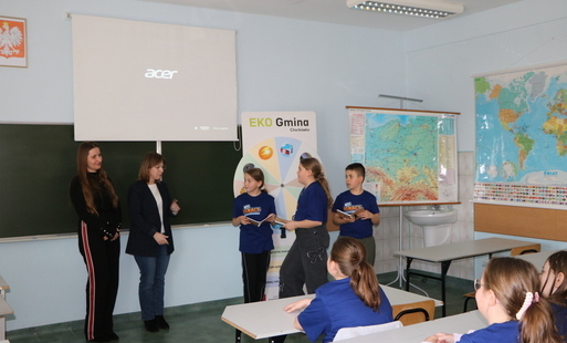 W sali lekcyjnej w Chorkówce kobieta w granatowej marynarce stoi po lewej stronie i trzyma w rękach kartki papieru. Po jej prawej stronie stoją troje dzieci. Dwie dziewczynki mają na sobie niebieskie koszulki z napisem "Być jak Ignacy". Chłopiec ma czarne