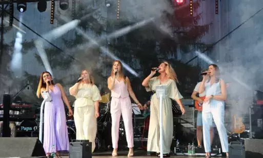 Pięć kobiet w pastelowych kombinezonach śpiewa na scenie, trzymając mikrofony. Za nimi widać muzyków grających na instrumentach. Oświetlenie sceniczne i sprzęt muzyczny tworzą tło.