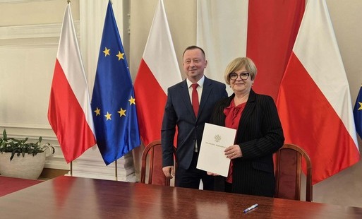 Szerokie ujęcie przedstawiające kobietę i mężczyznę stojących przy długim, ciemnym, drewnianym stole w oficjalnym pomieszczeniu. Mężczyzna po lewej ma na sobie ciemny garnitur i czerwony krawat, natomiast kobieta po prawej nosi czarną marynarkę w prążki i