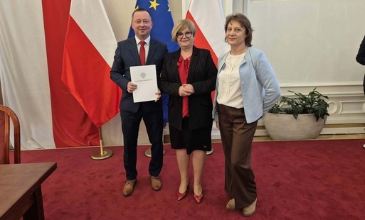Trzy osoby stojące obok siebie w oficjalnym wnętrzu na tle polskiej i unijnej flagi. Po lewej stronie stoi mężczyzna w garniturze, który trzyma białą teczkę z godłem Polski i napisem „Wojewoda Podkarpacki”. W środku stoi kobieta w czarnej marynarce i czer