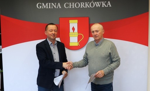 Dwaj mężczyźni, znani z poprzednich zdjęć, stoją przed oficjalną ścianką Gminy Chorkówka. Mężczyzna w marynarce (po lewej) i starszy mężczyzna w szarym swetrze (po prawej) wymieniają mocny uścisk dłoni. W drugich rękach trzymają egzemplarze podpisanej umo