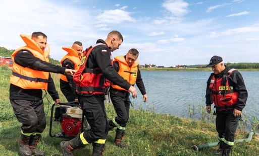 Pięciu strażaków nad brzegiem jeziora przenosi czerwoną motopompę spalinową. Część z nich ma na sobie pomarańczowe kamizelki ratunkowe, a inni czerwone kamizelki asekuracyjne.