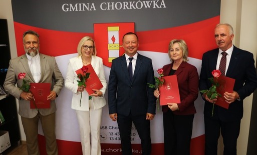 Fotografia przedstawia oficjalną grupę pięciu osób pozujących w pomieszczeniu na tle biało-czerwonej ścianki z napisem „GMINA CHORKÓWKA” i herbem przedstawiającym lampę naftową. W centrum stoi mężczyzna w ciemnym garniturze (Wójt). Po jego prawej stronie 
