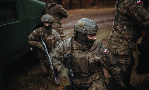 Scena taktyczna z udziałem czterech żołnierzy ubranych w mundury wz. 93 i hełmy z maskowaniem. Na pierwszym planie żołnierz w balaklawie patrzy w bok, a pozostali stoją w różnych pozach, trzymając broń długą. Po lewej stronie widać fragment zielonego poja