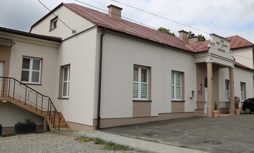 Frontowa część budynku z reprezentacyjnym wejściem głównym podpartym dwoma filarami (kolumnami). Nad wejściem widnieje napis „1936 DOM LUDOWY w ZRĘCINIE”. Po lewej stronie znajdują się schody zewnętrzne oraz wjazd/plac utwardzony.