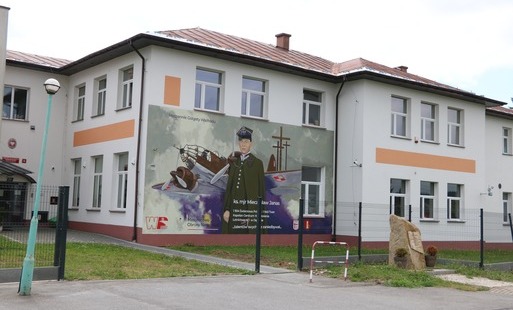 Zdjęcie przedstawiające budynek użyteczności publicznej (placówkę oświatową) w Świerzowej Polskiej, z widocznym nowym, panelowym ogrodzeniem na podmurówce modułowej. Na elewacji frontowej budynku znajduje się wielkoformatowy mural upamiętniający ks. mjr. 