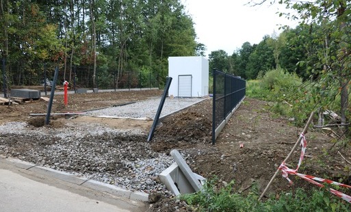 Zdjęcie przedstawia teren budowy kontenerowej pompowni wodociągowej przy drodze asfaltowej. Na ogrodzonej, utwardzonej żwirem działce stoi biały, prostopadłościenny budynek pompowni. Obok widoczny jest czerwony hydrant, słupki pod nowe ogrodzenie oraz bet