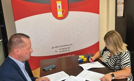 Zdjęcie wykonane z boku i z góry przedstawia dwie osoby przy biurku w pomieszczeniu biurowym. Mężczyzna o krótkich włosach, ubrany w marynarkę i koszulę w kropki, siedzi po lewej stronie, trzymając długopis nad kartką. Kobieta z blond włosami, w pasiastej