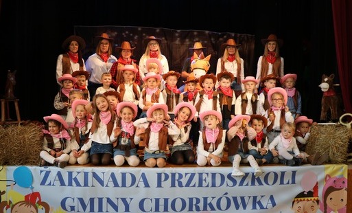 Zdjęcie grupowe pzredszkolaków, dziewczynki w różowych kalepuszach, chłopcy w brązowych. Panie wychowawczynie z przedszkola także w stylizacjach kowbojskich. Pozują do grupowego zdjęcia na scenie.  