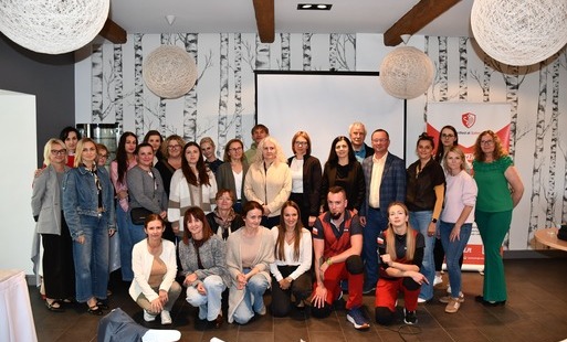 Zdjęcie grupowe wszystkich uczestników szkolenia oraz instruktorów, wykonane na koniec wydarzenia. Wszyscy stoją i klęczą, uśmiechnięci, patrząc w obiektyw aparatu. Tło stanowią charakterystyczna tapeta w brzozy, ekran projektora i baner reklamowy. Uczestnicy są w różnym wieku, ubrani w stroje codzienne. Zdjęcie upamiętnia zakończone szkolenie.