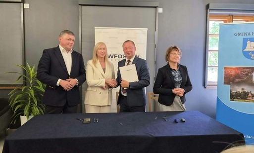 Na tym zdjęciu cztery osoby stoją. Pośrodku mężczyzna w garniturze i kobieta w jasnym kostiumie stoją naprzeciwko siebie, uśmiechają się i podają sobie dłonie, co sugeruje finalizację umowy. Mężczyzna trzyma w drugiej ręce teczkę z dokumentami. Po ich bokach, również stojąc, znajdują się pozostali uczestnicy spotkania: po lewej mężczyzna w ciemnej marynarce, a po prawej kobieta w okularach i ciemnej bluzce. Stół z obrusem i leżącymi na nim przedmiotami oraz oba roll-upy są nadal widoczne w tle.