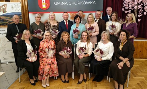 Grupa kilkunastu dyrektorów i przedstawicieli placówek oświatowych pozuje do pamiątkowej fotografii na tle ścianki promocyjnej „Gmina Chorkówka”. Osoby z pierwszego rzędu siedzą na krzesłach, a za nimi stoi drugi rząd uczestników spotkania. Prawie wszyscy obecni trzymają w rękach dekoracyjne kompozycje z ozdobnych wrzosów w doniczkach zawiniętych w papier. W tle po prawej stronie widoczne jest ozdobne drzewko z różowymi kwiatami.