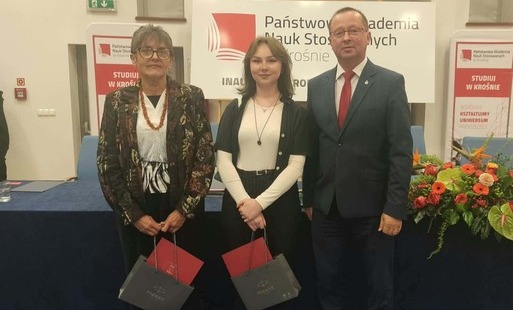 Zdjęcie pozowane wykonane w kuluarach lub w bocznej części auli. Trzy osoby stoją na tle ścianek reklamowych Państwowej Akademii Nauk Stosowanych w Krośnie z hasłami „STUDIUJ W KROŚNIE”. Od lewej: starsza kobieta w wzorzystej marynarce i naszyjniku, młoda kobieta w białej bluzce i czarnej narzutce (wyróżniona absolwentka) oraz mężczyzna w ciemnym garniturze i czerwonym krawacie. Obie kobiety trzymają czerwone dyplomy/listy gratulacyjne oraz ciemne torby upominkowe z logo „Parker”. Przed nimi widać fragment dekoracji kwiatowej.