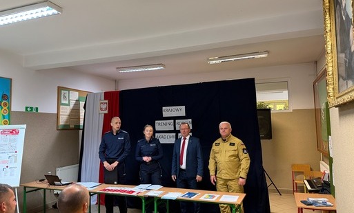 Czworo prelegentów stoi w szeregu za stołami z ulotkami informacyjnymi. Od lewej: policjant z czarną teczką, policjantka, mężczyzna w garniturze i czerwonym krawacie oraz mężczyzna w piaskowym mundurze. W tle wisi ciemna przesłona z białymi napisami oraz flaga Polski z godłem.