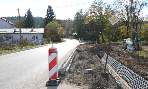 Prace budowlane przy drodze powiatowej polegające na budowie nowego chodnika oraz profilowaniu i umacnianiu rowów odwadniających. Na zdjęciach widoczne są odcinki drogi z ustawionymi biało-czerwonymi pachołkami ostrzegawczymi, wykopana i wyrównana ziemia pod krawężniki i podbudowę, a także fragmenty rowów wyłożone ażurowymi płytami betonowymi. W tle widoczne są domy jednorodzinne, zielone oraz jesienne drzewa i znaki drogowe.