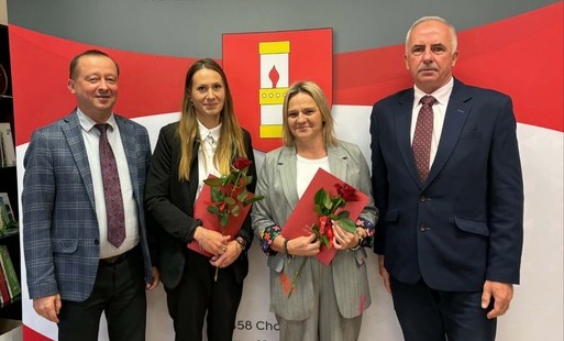 Fotografia przedstawia oficjalne pamiątkowe zdjęcie uczestników uroczystości na tle ścianki z napisem „GMINA CHORKÓWKA” oraz herbem gminy (przedstawiającym żółtą lampę naftową na czerwonym tle).   W centralnej części stoją dwie nauczycielki, które złożyły uroczyste ślubowanie – obie trzymają w dłoniach czerwone teczki z dokumentami awansu oraz po jednej czerwonej róży. Po ich bokach stoją dwaj mężczyźni w garniturach i krawatach (w tym Wójt Gminy Chorkówka, Grzegorz Węgrzynowski). Wszyscy uczestnicy uśmiechają się delikatnie do obiektywu, a kadr ma charakter pamiątkowy i oficjalny.