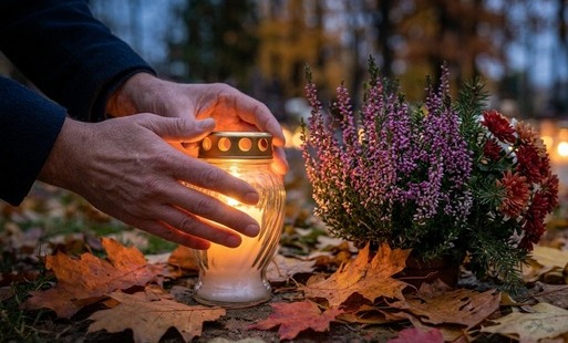 Jesienne ujęcie w odcieniach szarości i brązu: dłonie w ciemnym płaszczu osłaniają płomień znicza stojącego na ziemi wśród opadłych liści. Obok znicza znajduje się doniczka z fioletowymi wrzosami oraz bukiet jesiennych kwiatów. W tle rozmyte, ciepłe światła innych zniczy i drzewa w lekkiej mglistej poświacie.